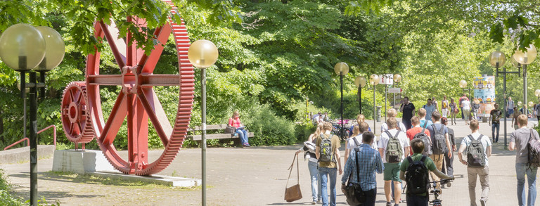 Kopfbild Studierende rote Zahnräder Campus Nord Studierende im Sommer auf dem Campus Nord, rote Zahnräder im Hintergrund